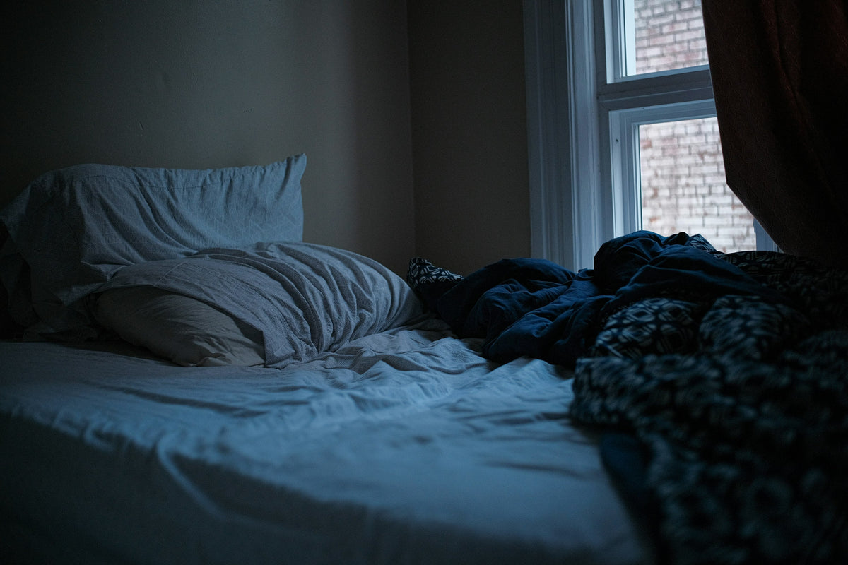 Unmade bed with white sheets and a patterned blanket beside a window