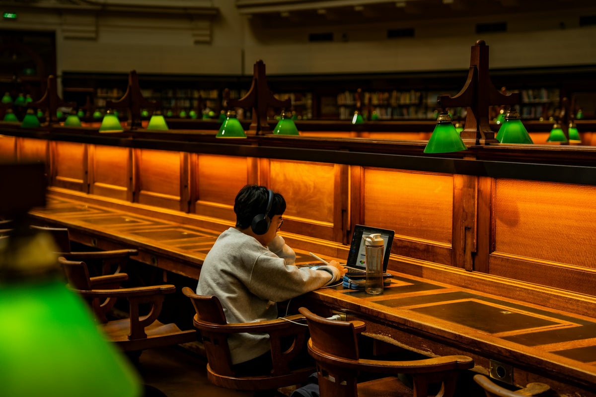 A student in headphones works on a laptop at a long wooden desk in a library, under reading lamps