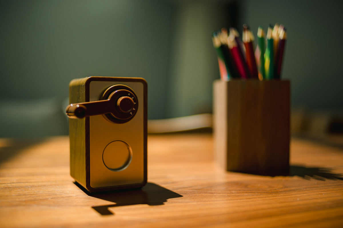 A hand-crank pencil sharpener and a wooden pot of coloured pencils on a wooden desk in low sunlight