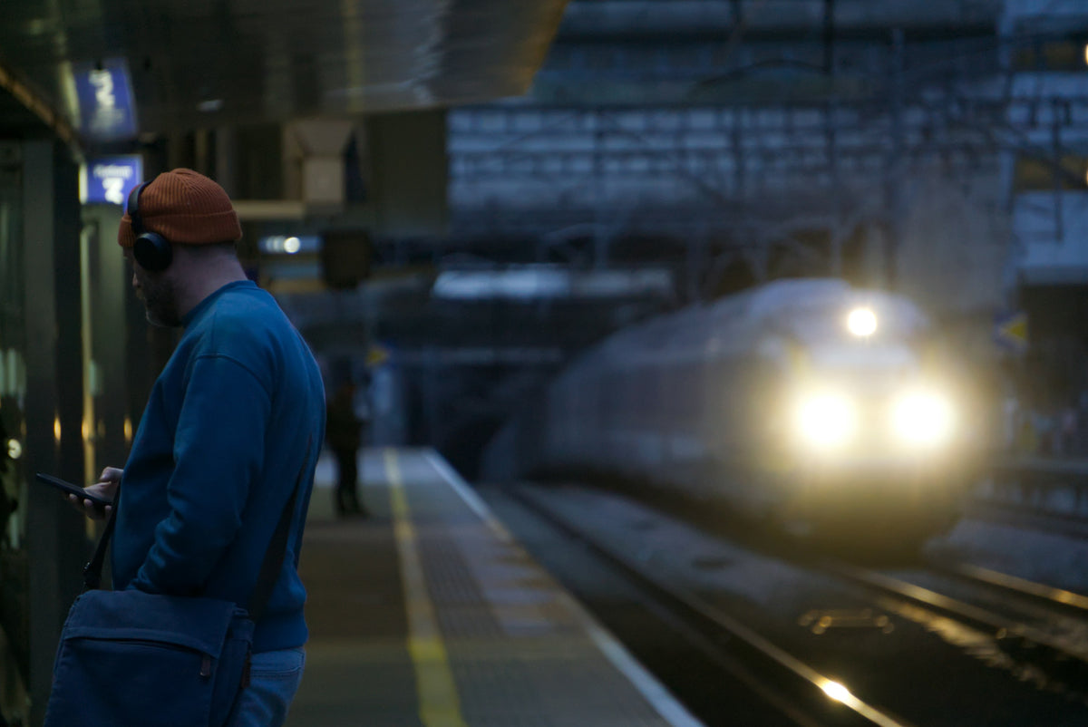 A man in headphones checks his phone on a station platform as a train comes in at dusk