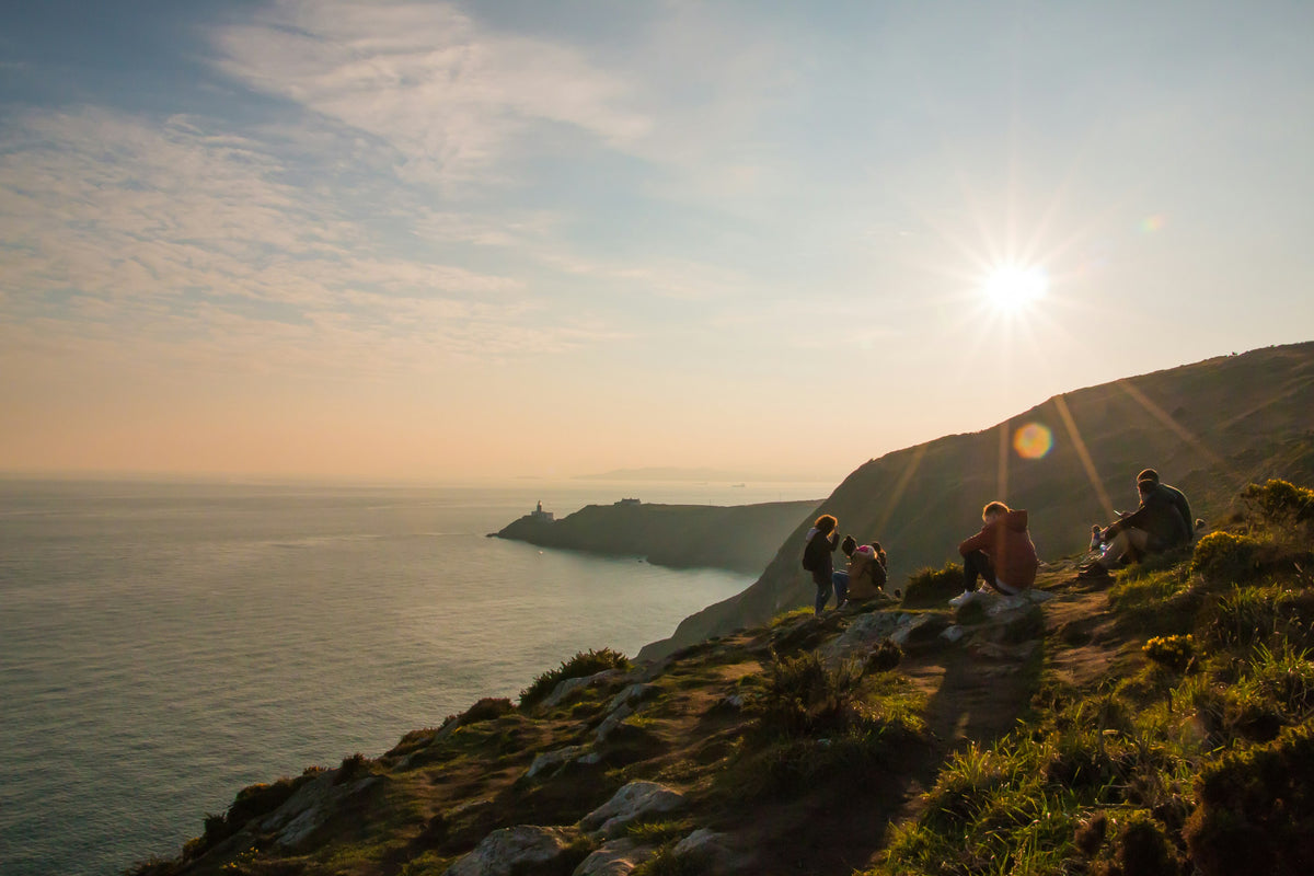 People sit on the grassy cliff path at Howth Head at sunset, with the Baily lighthouse and the sea behind