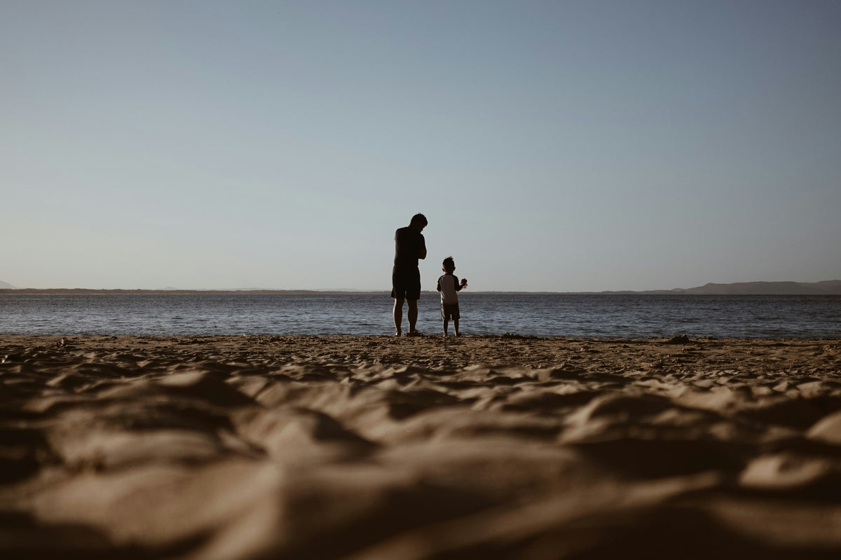 An adult and a child in silhouette at the edge of the sea, seen from low on a sandy beach