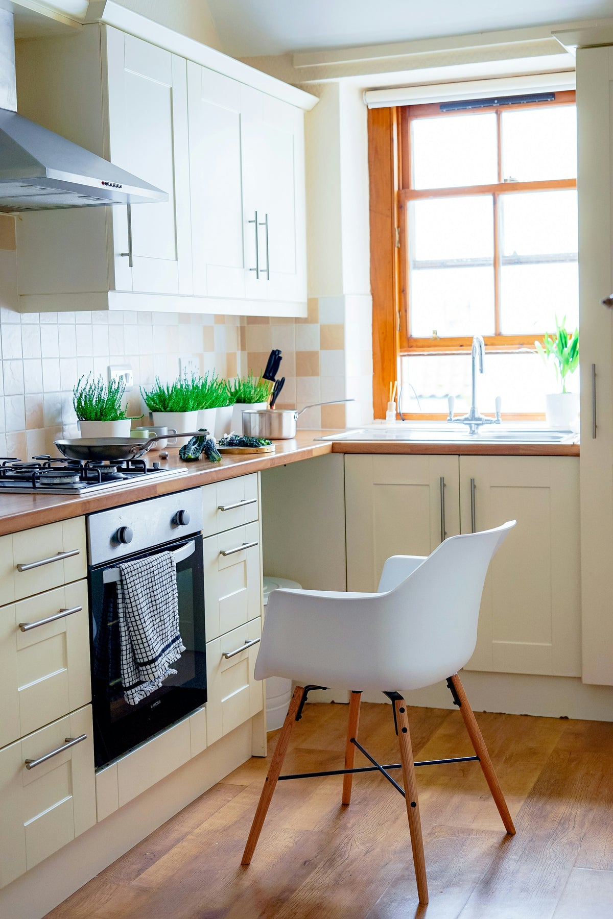 A bright kitchen with a hob and oven, cream units and a white chair by the window