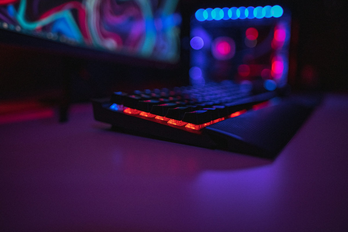 A backlit mechanical keyboard on a desk at night, with a monitor and PC lit up behind it