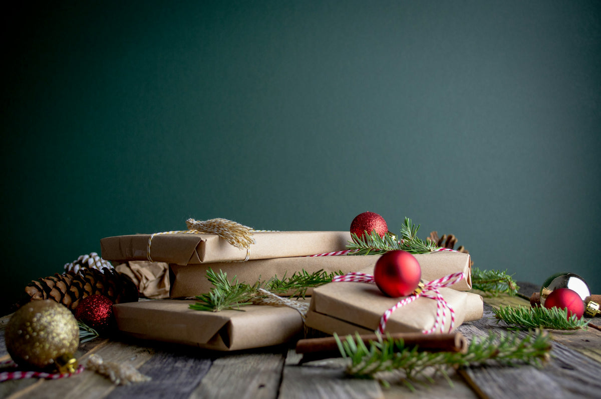 Presents wrapped in brown paper with red baubles and pine sprigs on a wooden table, in front of a dark teal wall