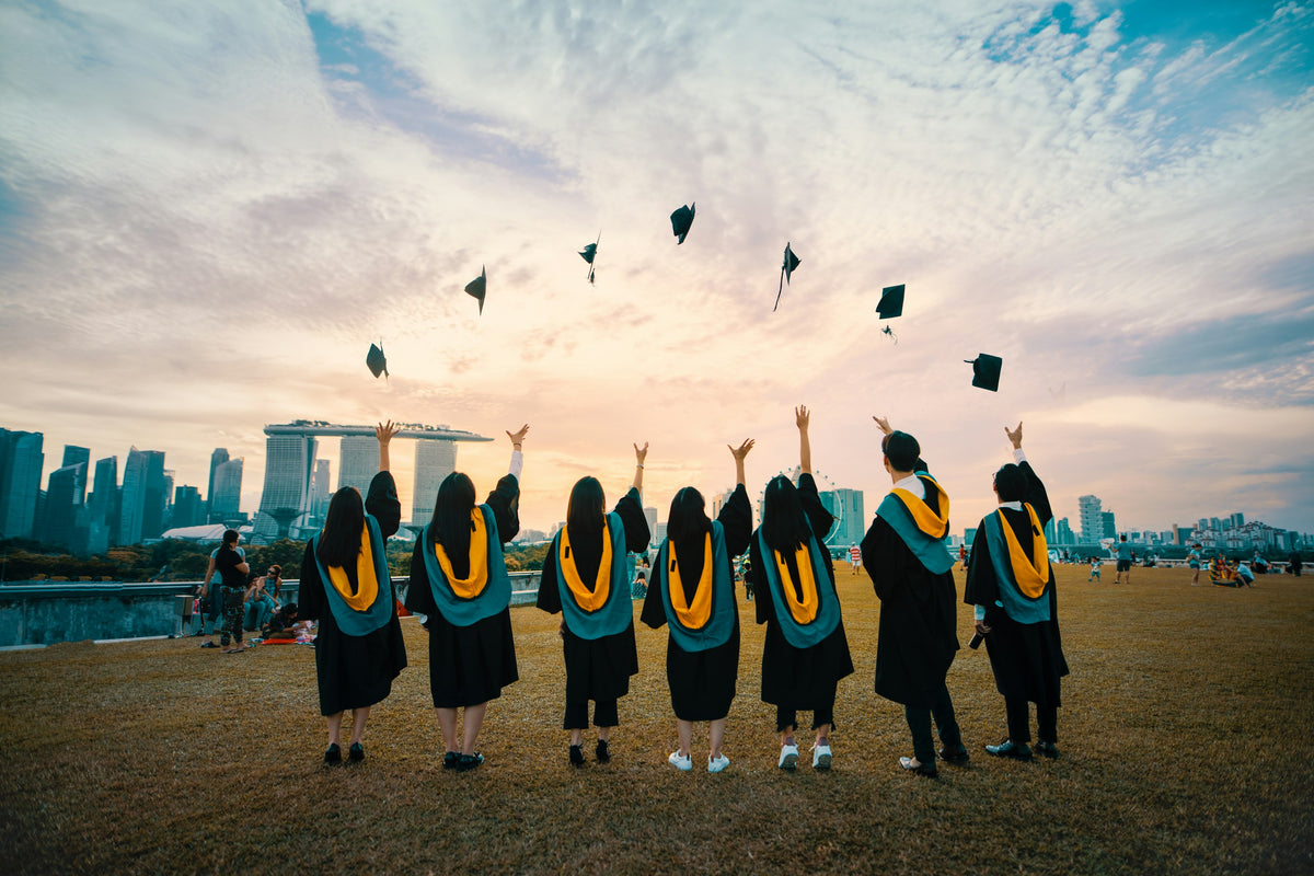 Graduates in gowns, seen from behind, throwing their caps into an evening sky