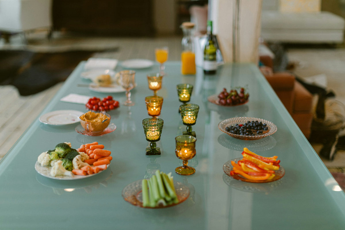 A glass table set with plates of vegetables, cherries and blueberries, glasses of juice and lit candles