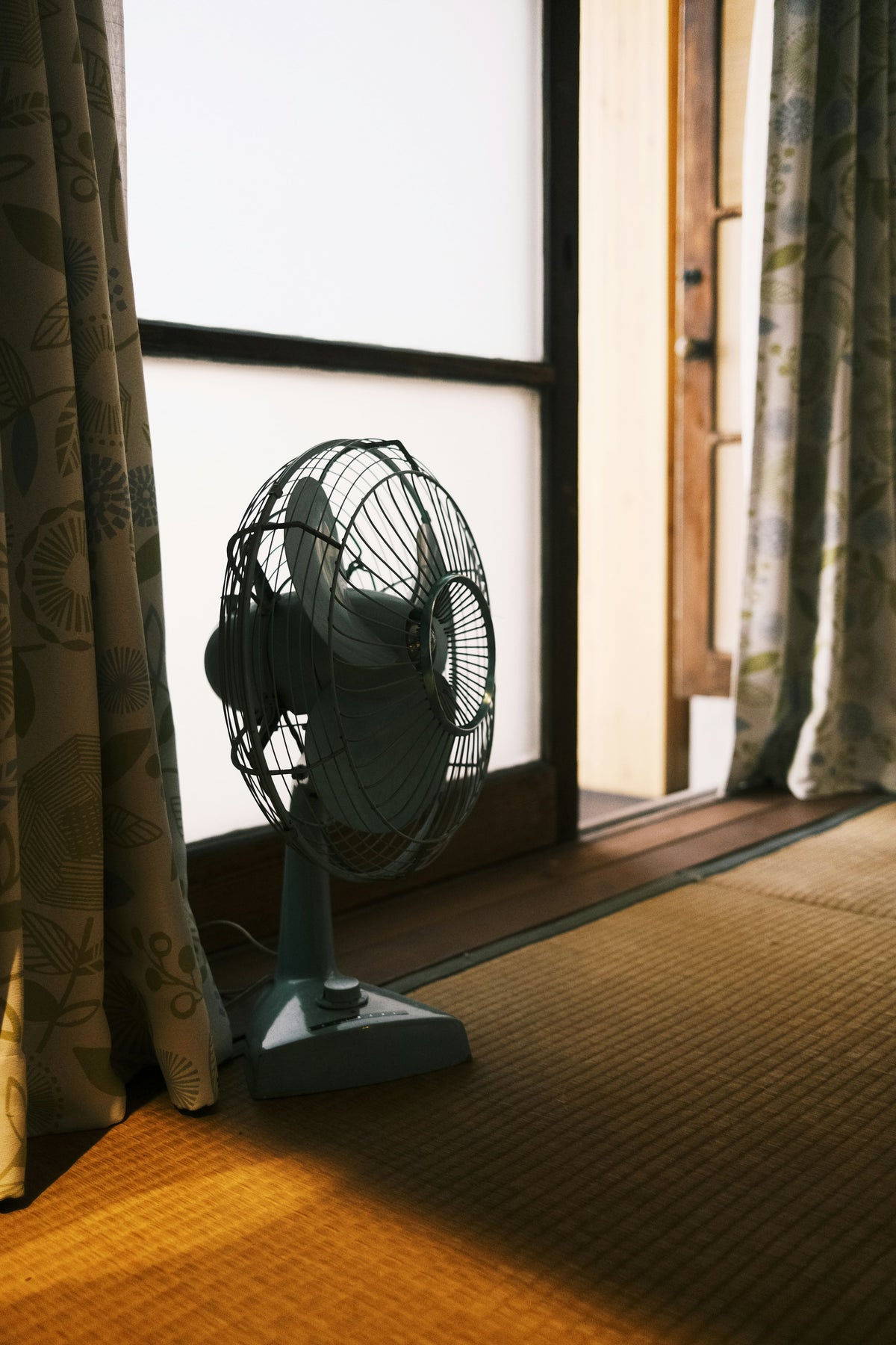 A desk fan on a wooden floor beside a sunny window with patterned curtains