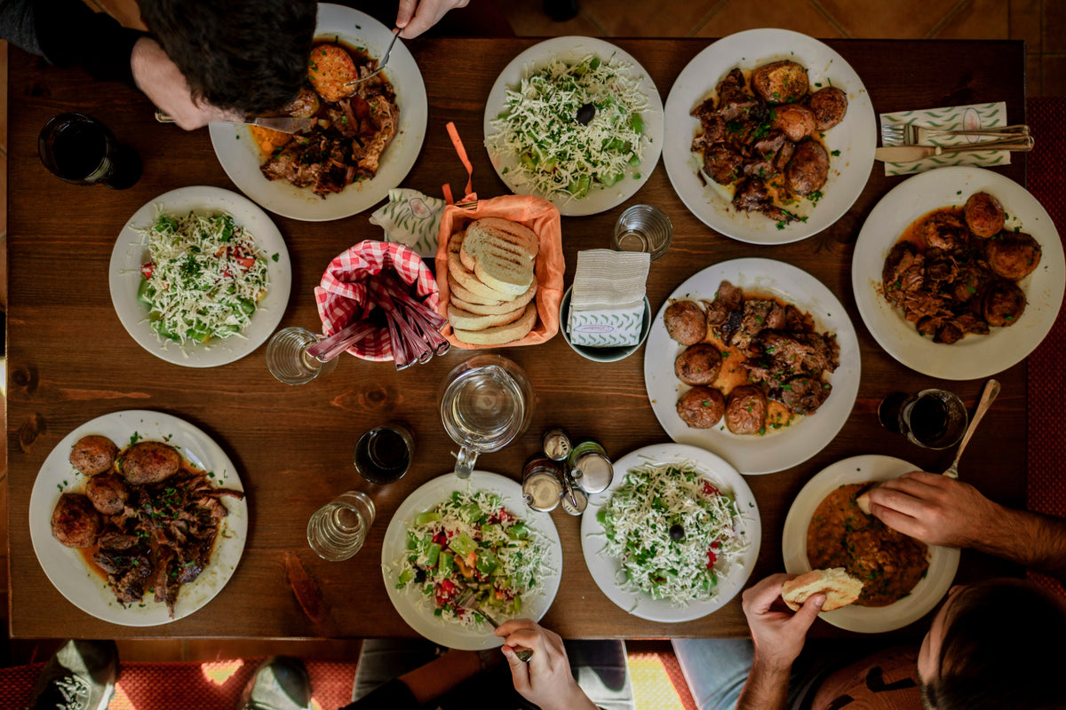 A wooden table seen from above, laid with a dozen plates of food, bread and water glasses
