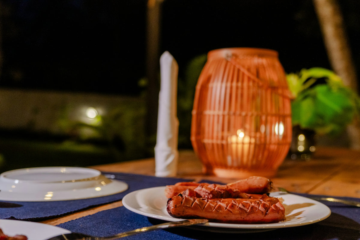 Evening table outdoors with a glowing lantern and grilled sausages on a plate