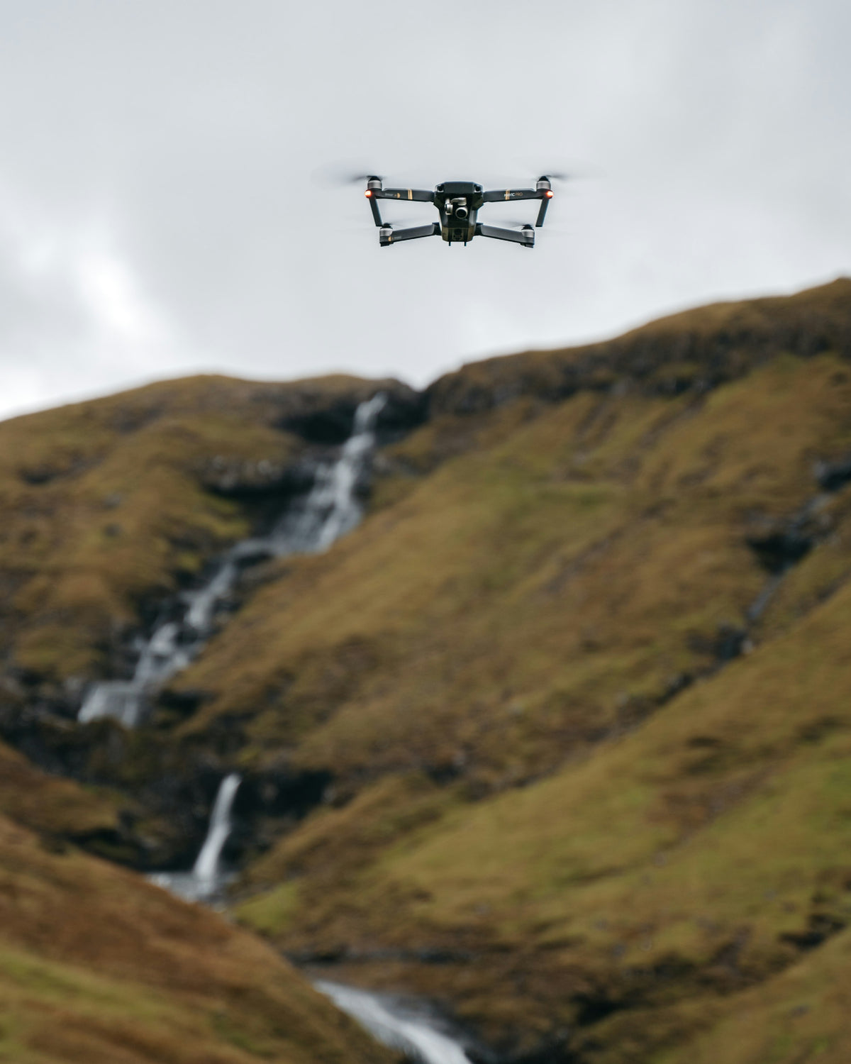 Grey camera drone in the air above a grassy valley with a waterfall