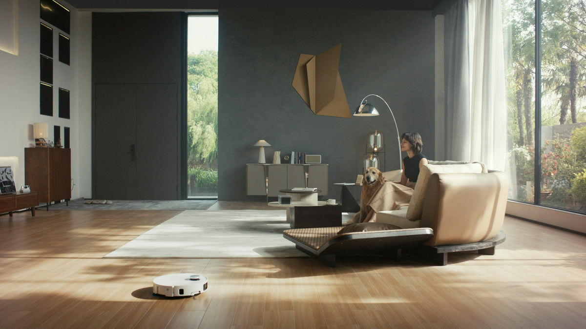 A white robot vacuum on a wooden floor in a bright living room, with a woman and a dog on the sofa