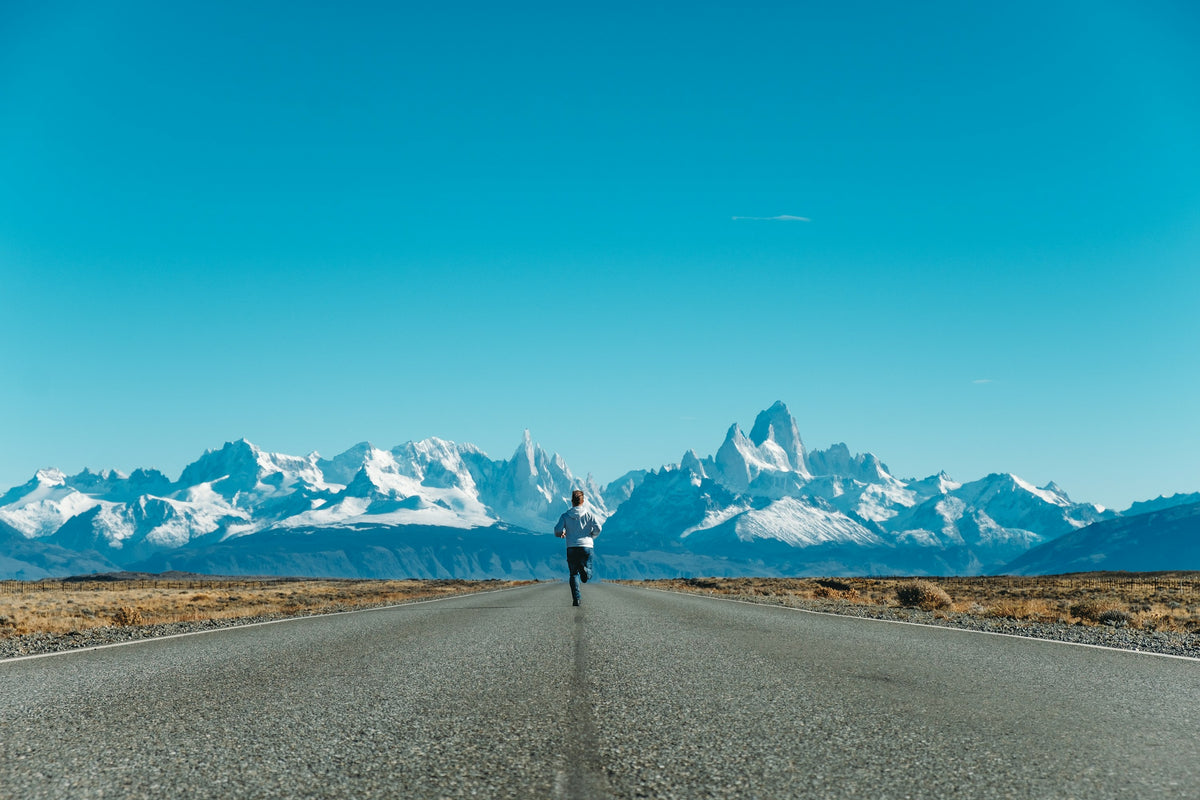 A runner on a long straight road towards snow-capped mountains under a blue sky