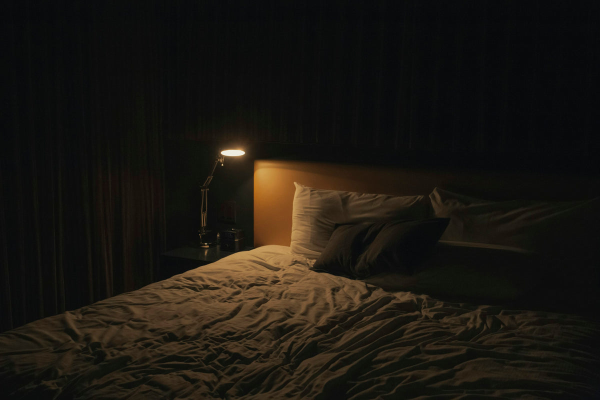 Dark bedroom with a made bed and a small lamp lit on the bedside table