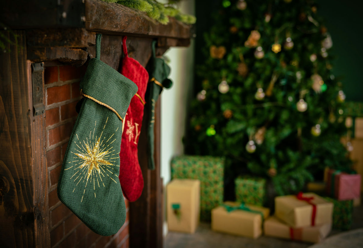 A green stocking with a gold star hanging from a wooden mantelpiece, with a Christmas tree and wrapped presents behind