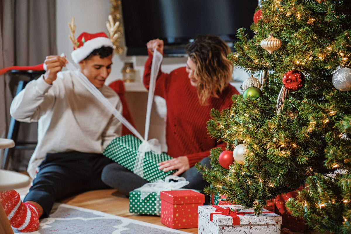 Two people opening wrapped presents beside a Christmas tree