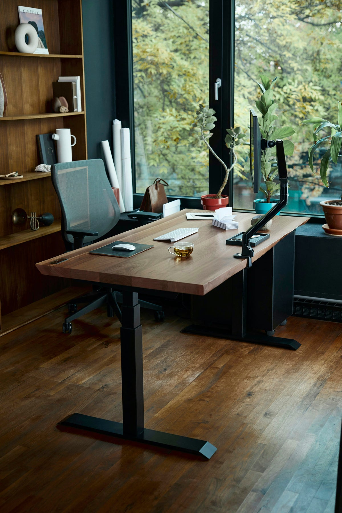 A wooden desk by a window with a monitor on a clamp-on arm, a keyboard, a mouse and an office chair